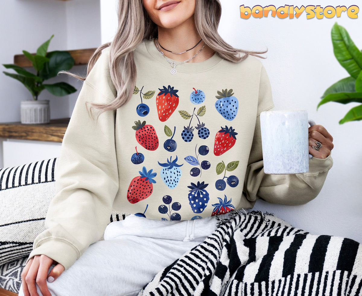 Cute Berry Sweatshirt in sand on seated person holding mug beside plants and striped textiles.