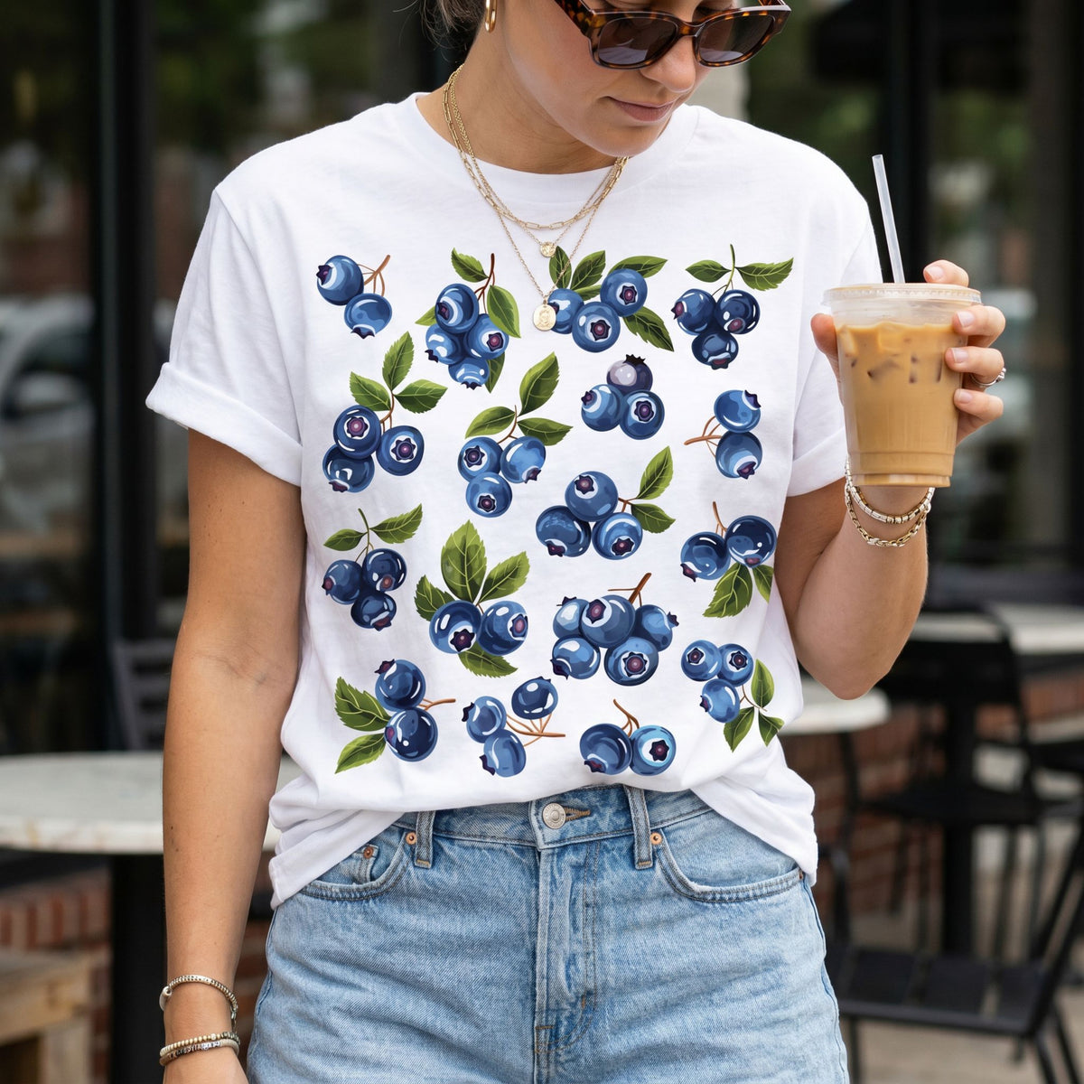 Cute Blueberry T-Shirt in white, front view outdoors with iced coffee, jeans, and patio seating.