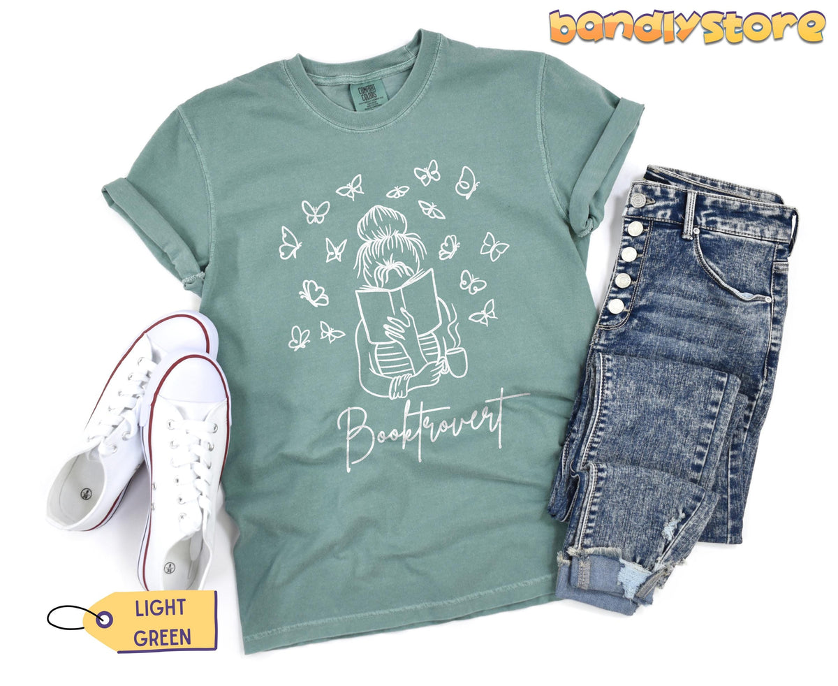 Booktrovert T-Shirt in light green flat-laid beside white sneakers and blue jeans.
