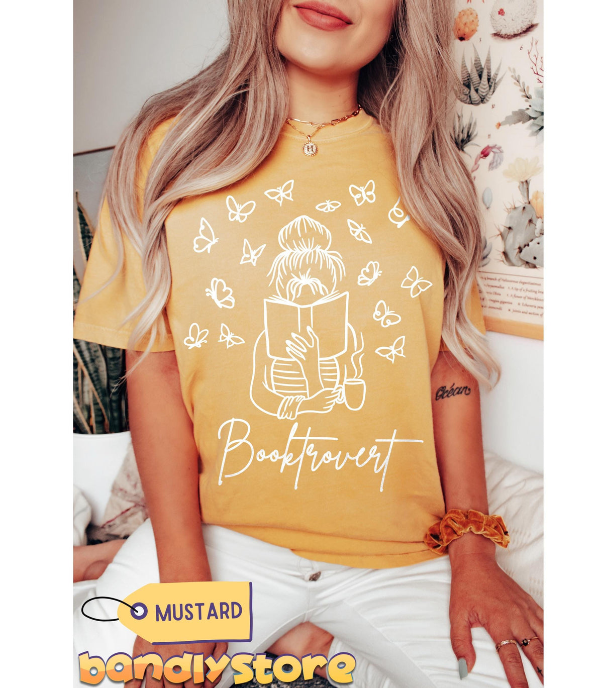 Booktrovert T-Shirt in mustard on a seated model shown front-on amid plants and botanical wall prints.