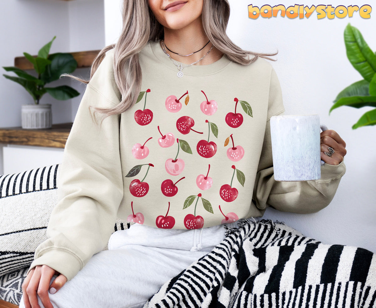 Cute Cherry Sweatshirt in sand, worn front-facing by a seated woman holding a mug amid plants and striped textiles.