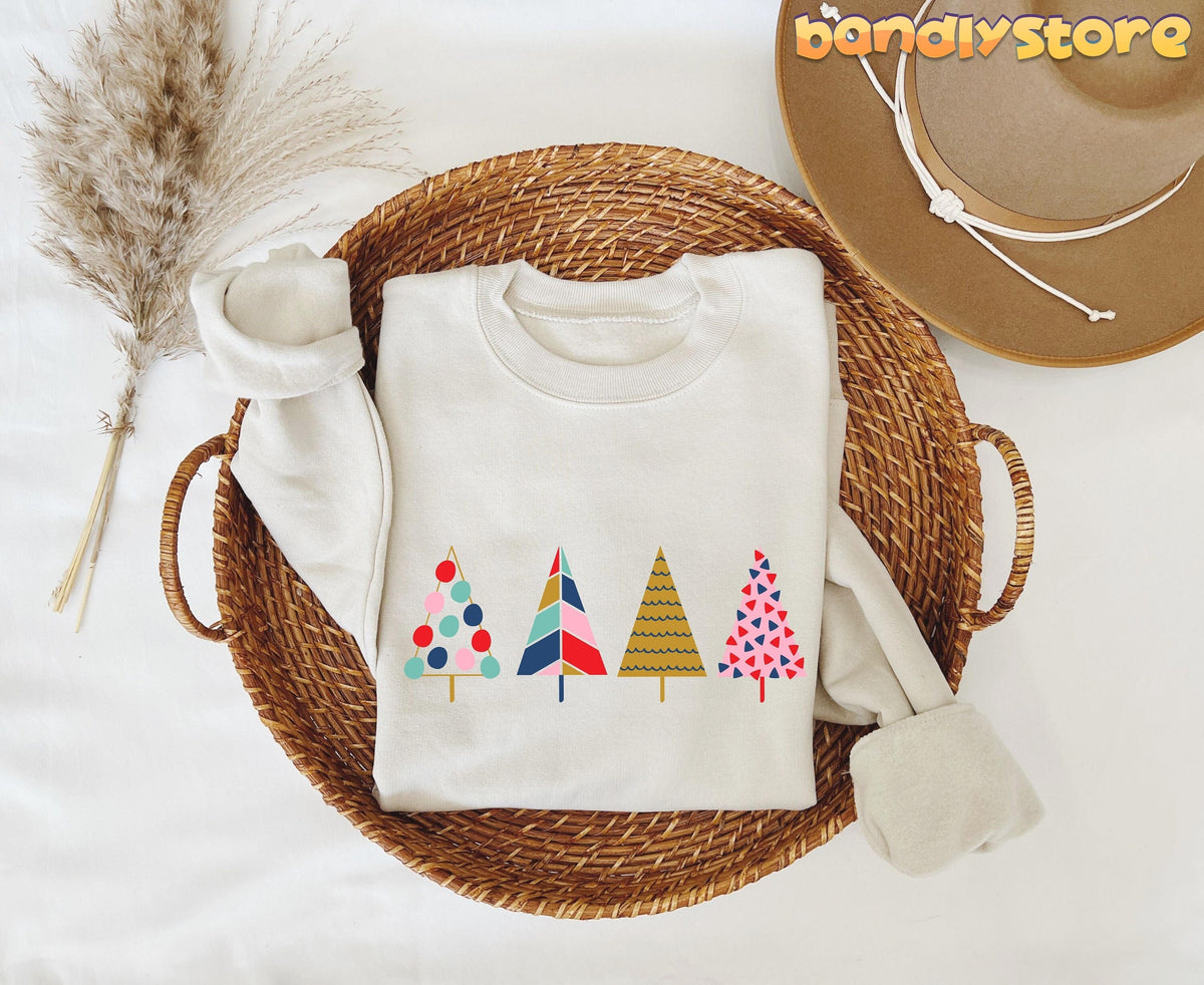 Cute Christmas Trees Sweatshirt in sand folded in a wicker basket beside pampas grass and a hat, top-down view.