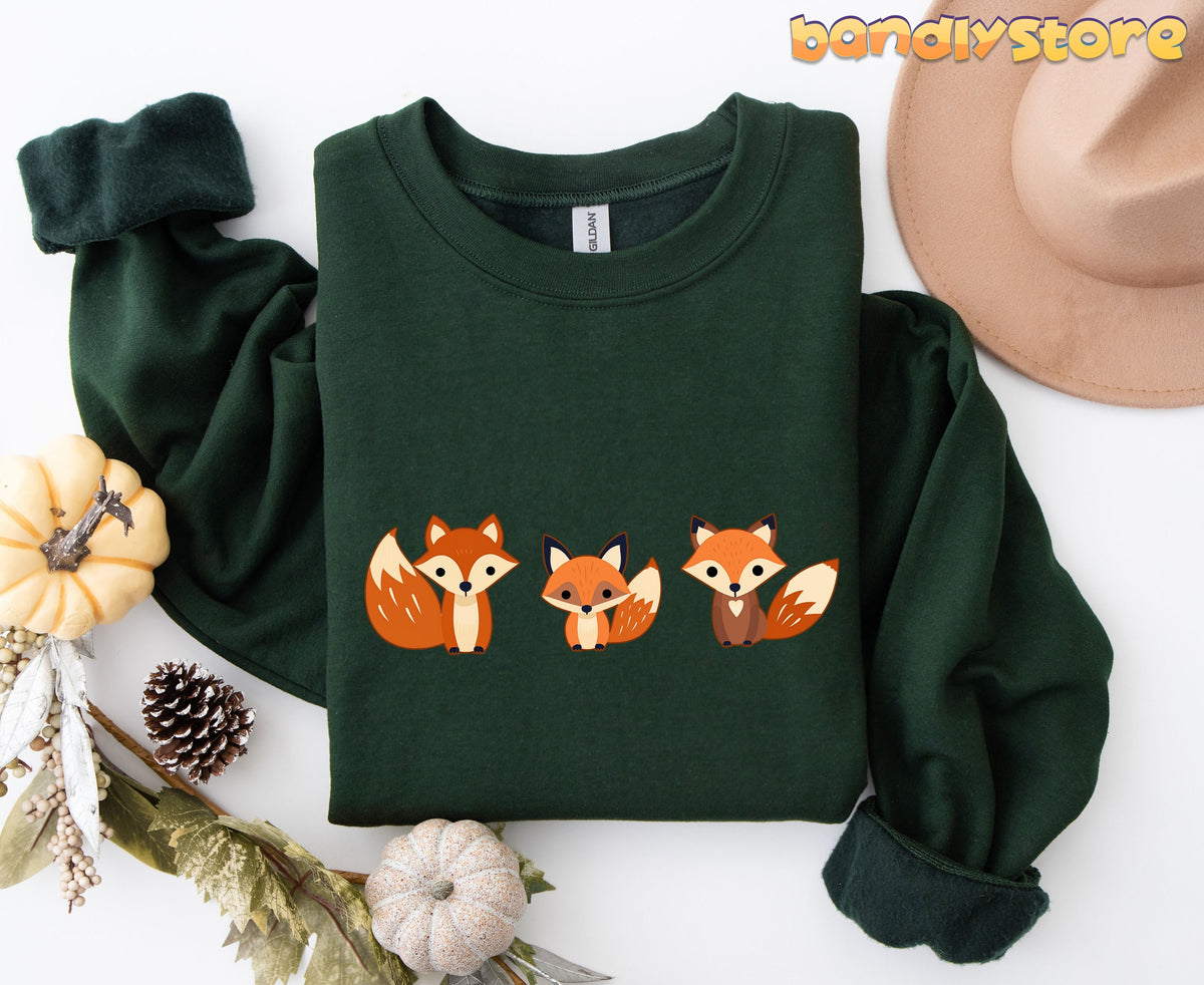 Cute Fox Sweatshirt forest green flat lay with three foxes beside pumpkins, hat, pinecone and leaves.