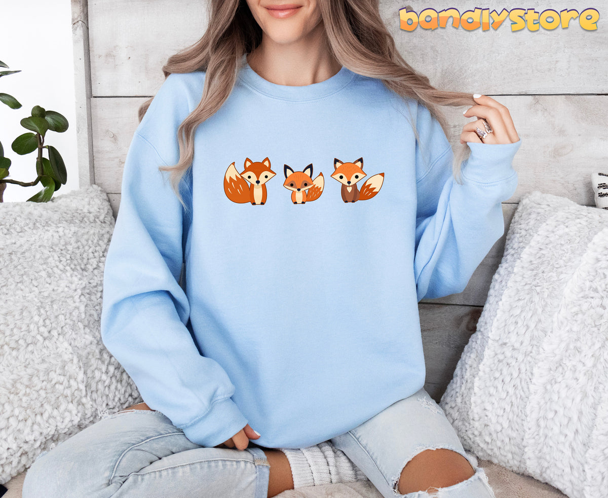 Cute Fox Sweatshirt in light blue, worn front-facing by a seated person amid pillows and a plant.