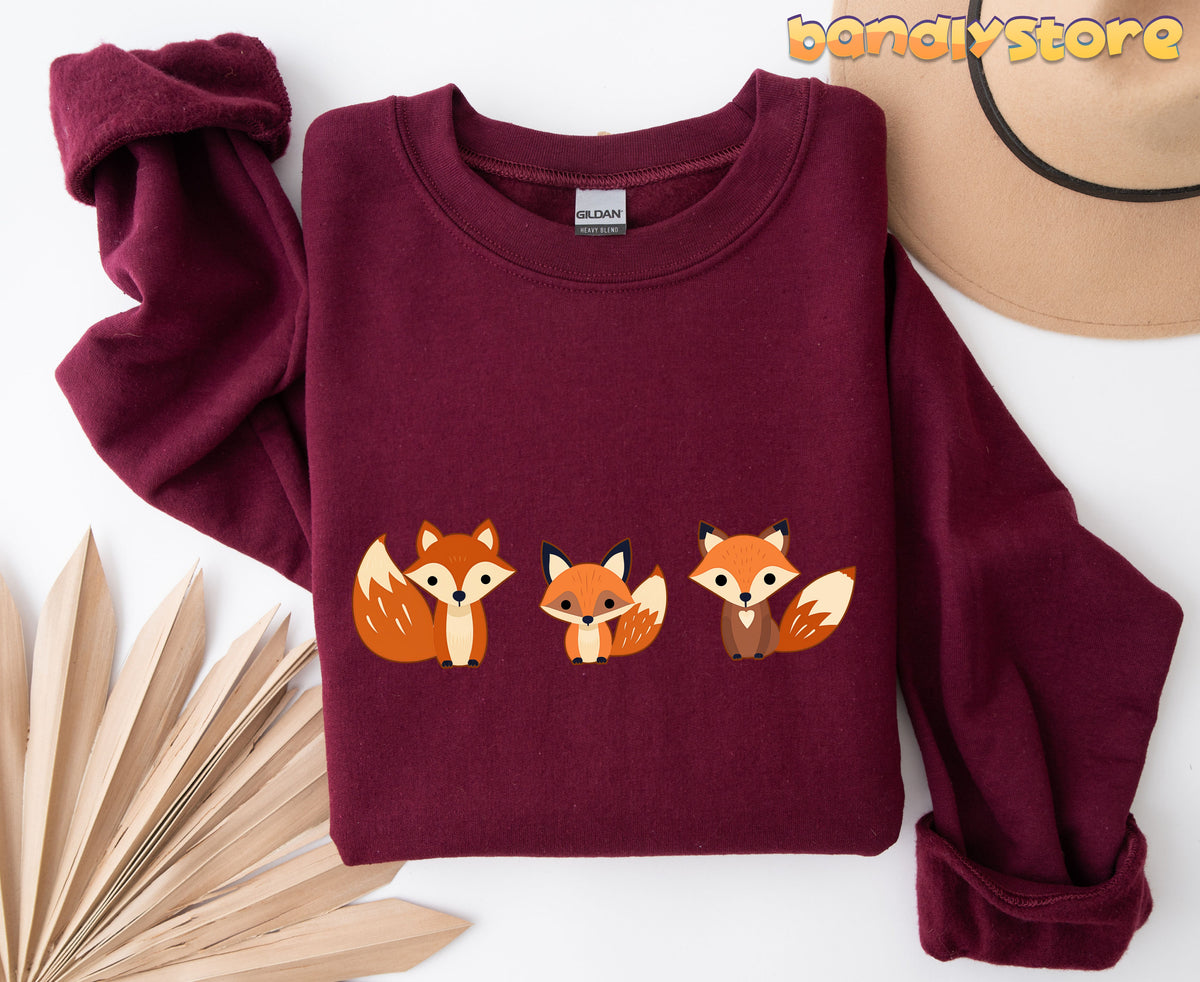 Cute Fox Sweatshirt in maroon folded flat from above beside a hat and dried leaves.