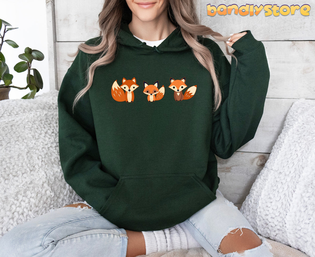 Cute Foxes Hoodie in Forest Green worn by a seated person on a fluffy white sofa beside a plant against wood panels.