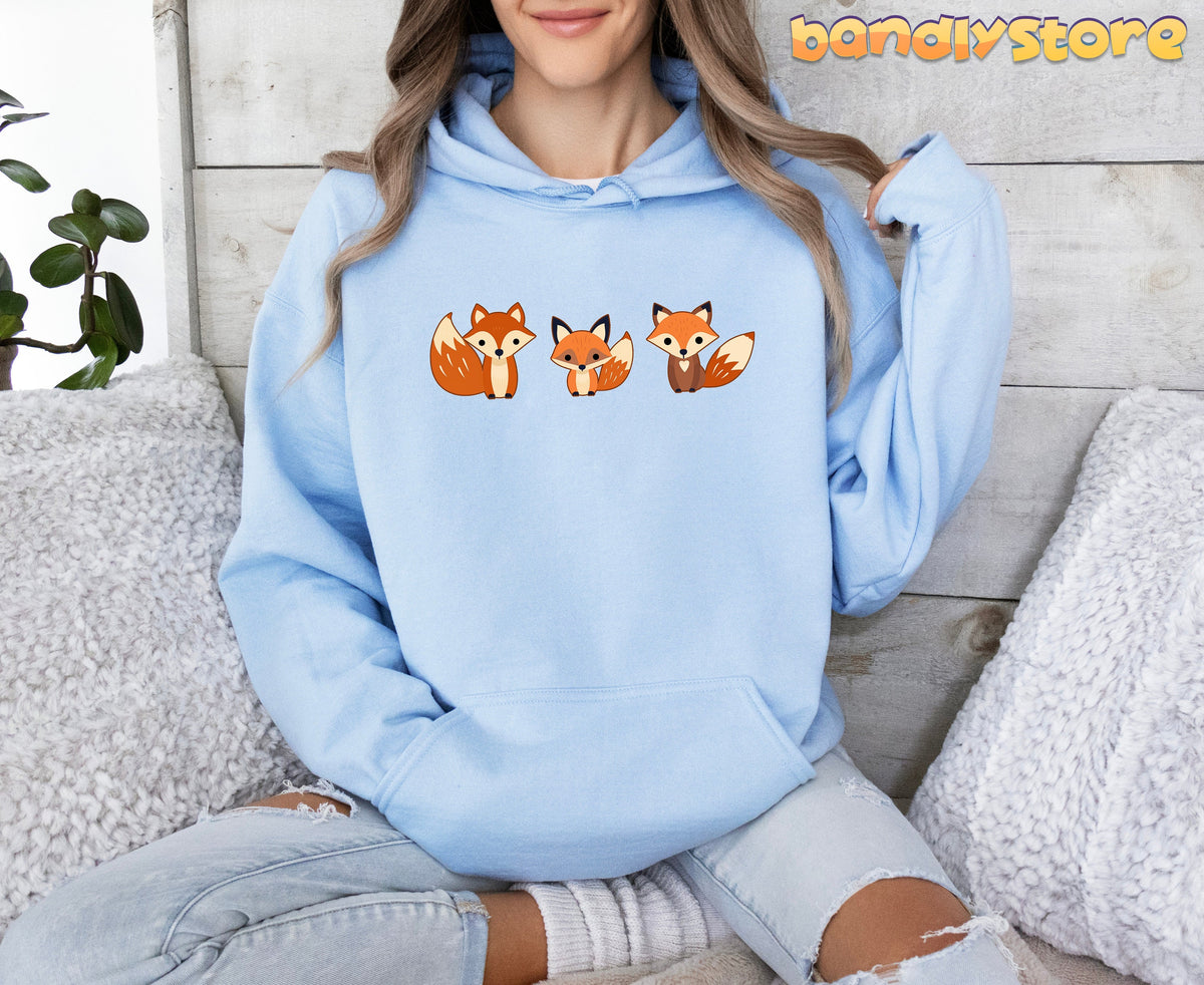 Cute Foxes Hoodie in light blue, front view on a seated model amid pillows, plant and wood wall.
