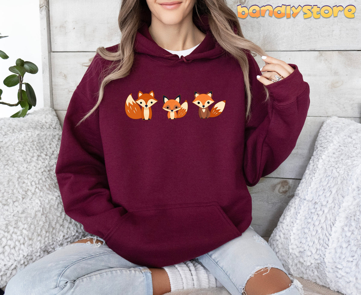 Cute Foxes Hoodie in maroon, worn by a seated person amid pillows and a plant against a wooden wall.