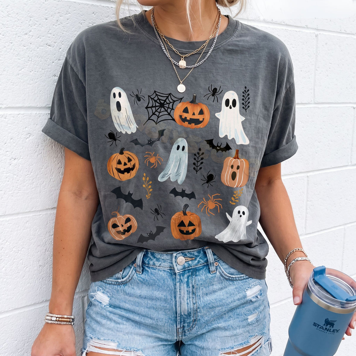 Cute Spooky Ghosts Pumpkins T-Shirt in pepper on a model against a white brick wall, front view, holding a tumbler.
