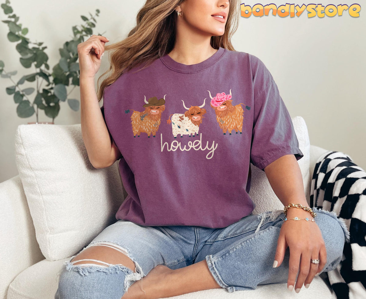 Cute Highland Cows T-Shirt in berry, front view on woman seated on white sofa beside plant and checkered blanket.