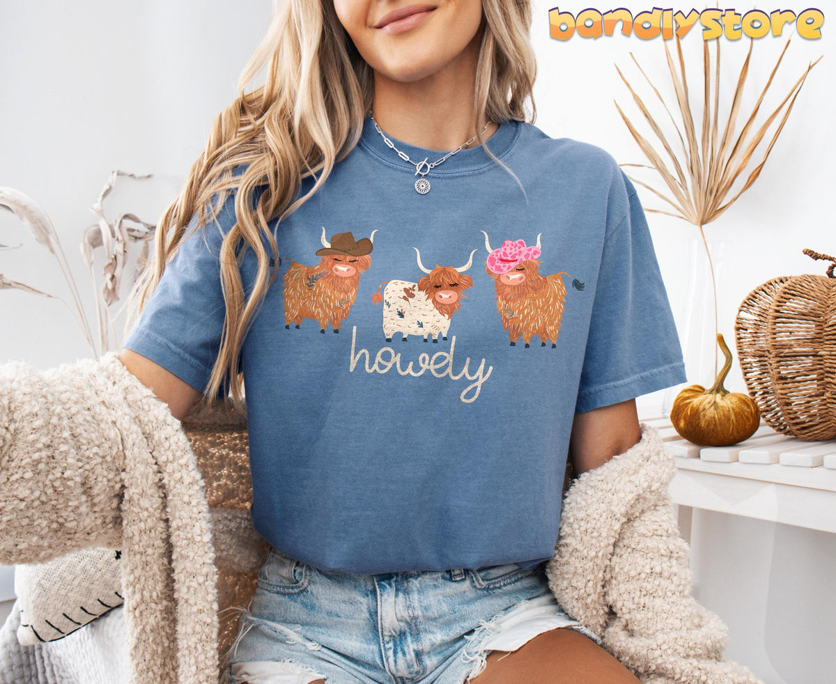 Cute Highland Cows T-Shirt in Blue Jean, worn by a seated woman amid dried plants, a pumpkin and woven décor.