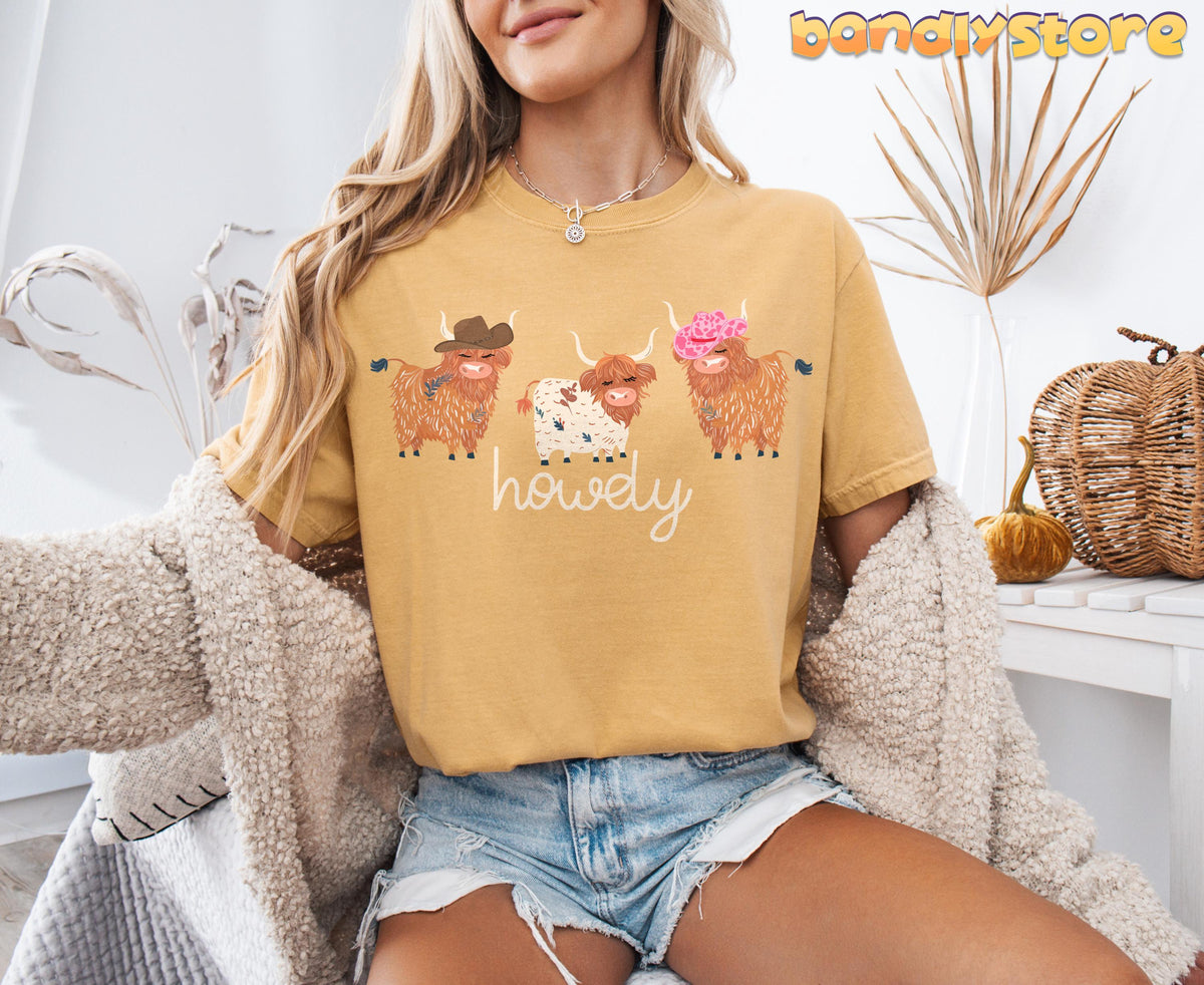 Cute Highland Cows T-Shirt in mustard worn front-facing by a seated woman beside dried plants and pumpkins.