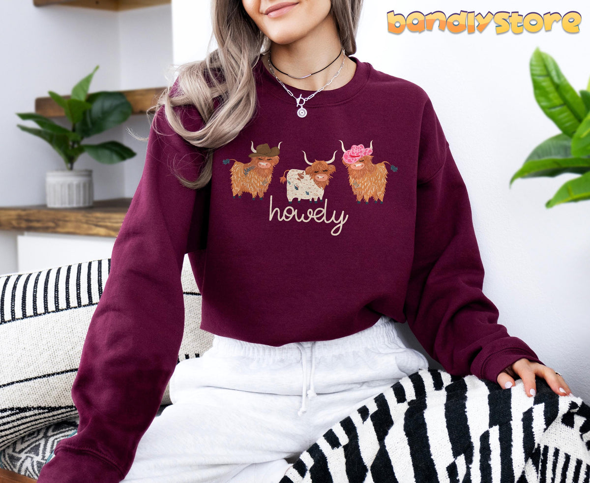 Cute Highland Cows Sweatshirt in maroon on a seated woman indoors beside plants and striped cushions.
