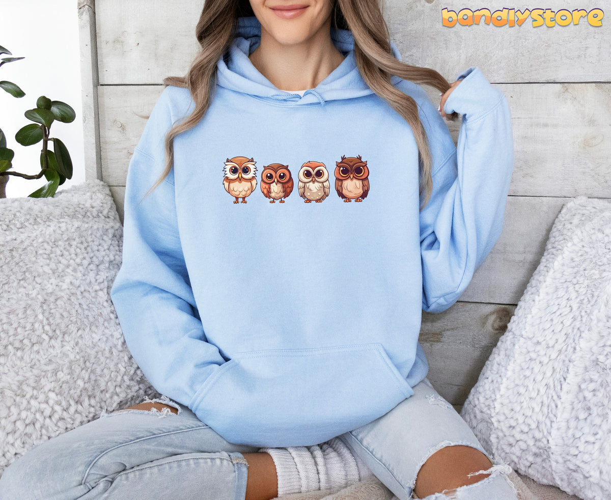 Cute Owl Hoodie in light blue on a seated person amid fluffy pillows and a plant against wood paneling.