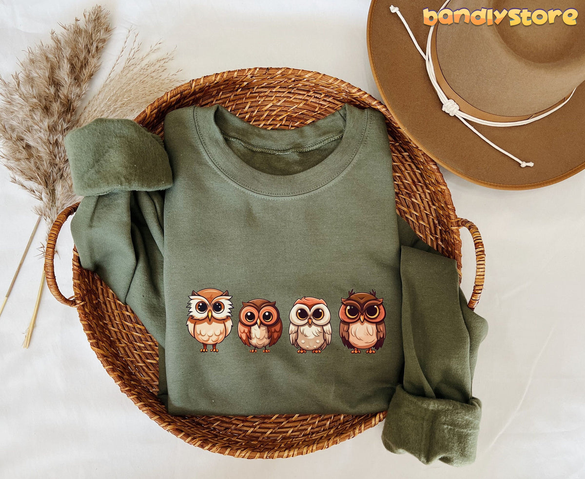 Cute Owl Sweatshirt in military green folded in a wicker basket beside pampas grass and a hat.