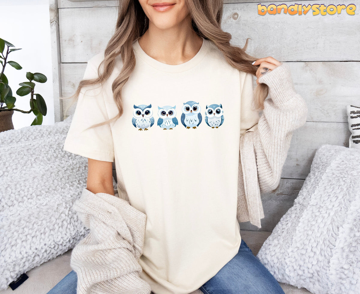 Cute Snow Owl T-Shirt in natural worn front-facing by a woman seated among pillows and a plant against a wooden wall.