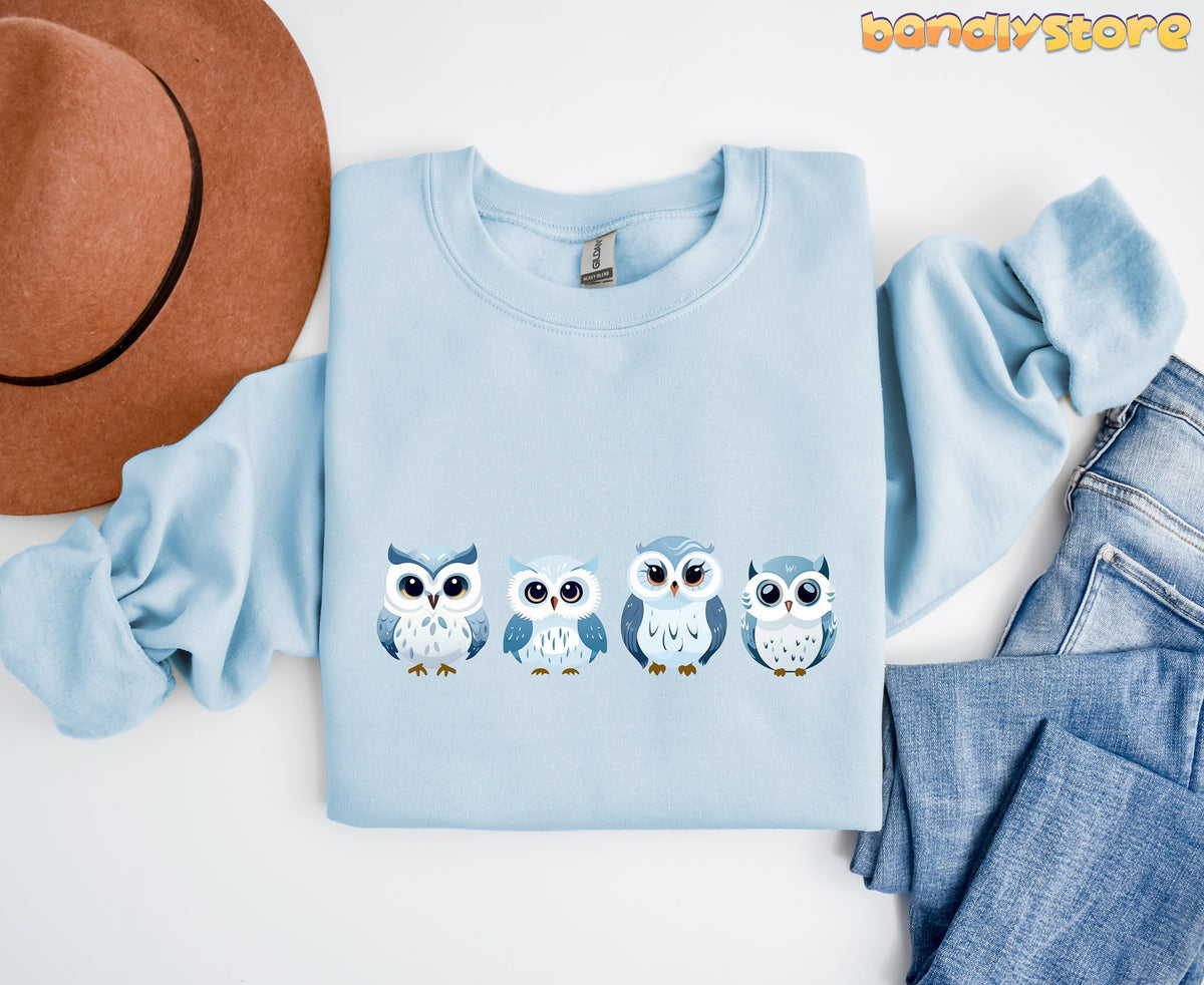 Cute Snow Owl Sweatshirt in light blue flat-laid from above beside a brown hat and jeans.