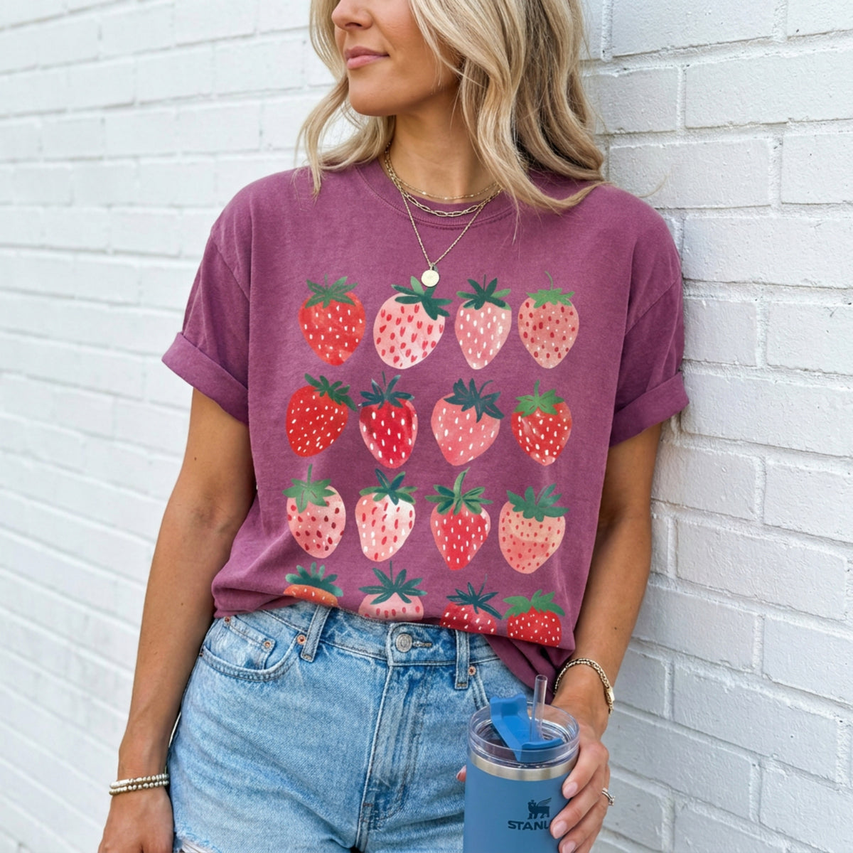 Cute Strawberry T-Shirt in berry on a woman leaning against a white brick wall holding a blue tumbler.
