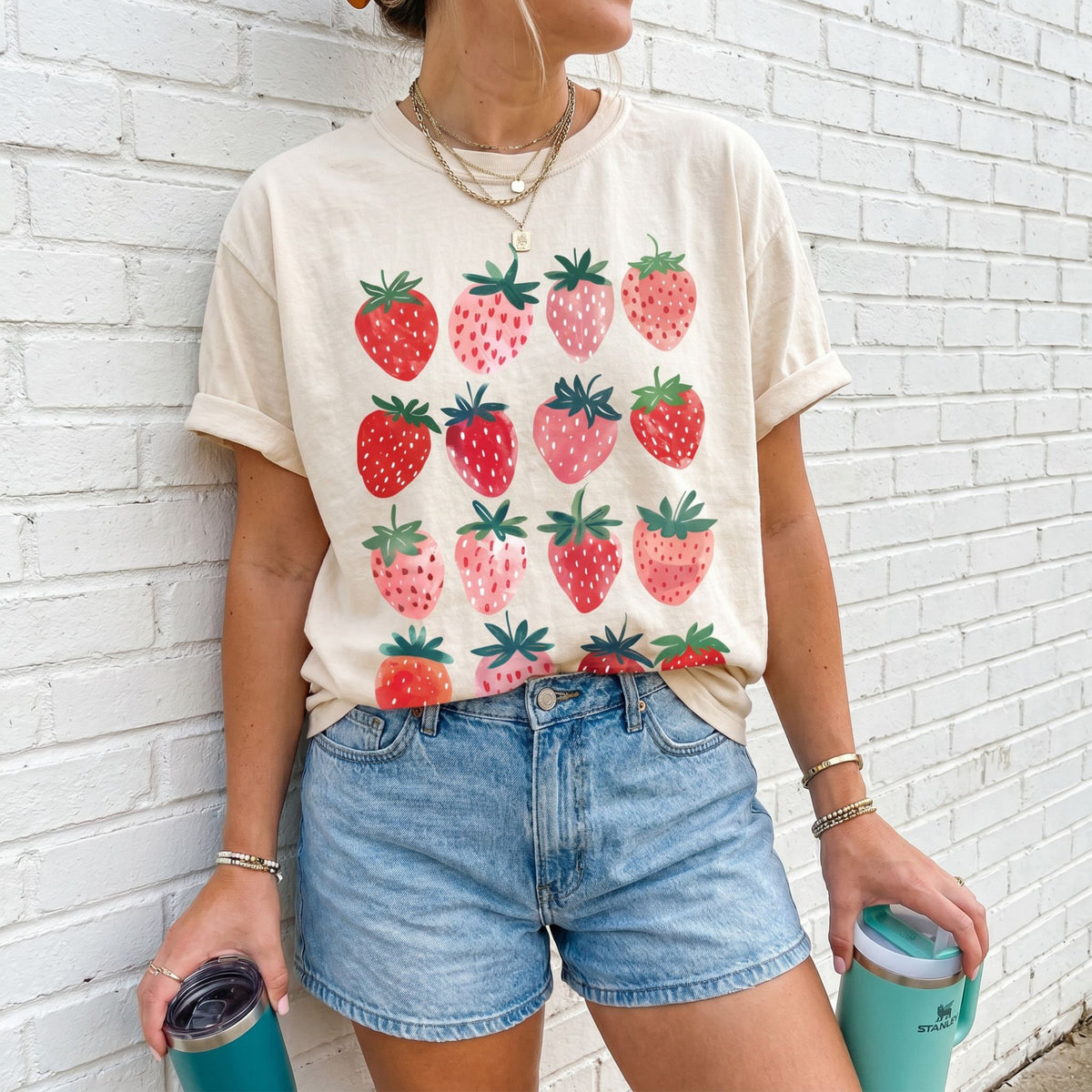 Cute Strawberry T-Shirt in ivory on a person against a white brick wall holding two tumblers.