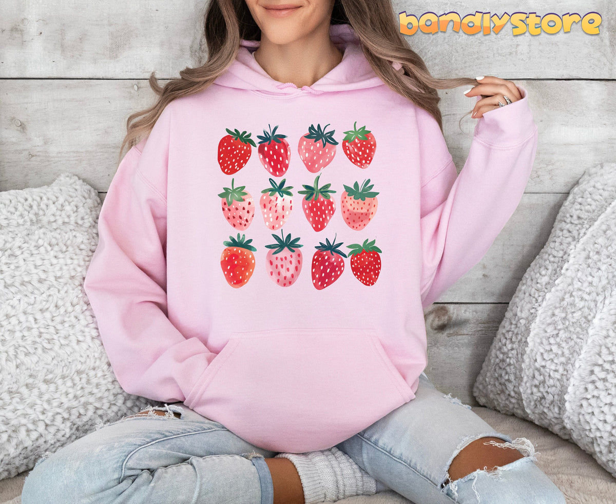 Cute Strawberry Hoodie in light pink, front view on seated model amid white pillows and wood wall.