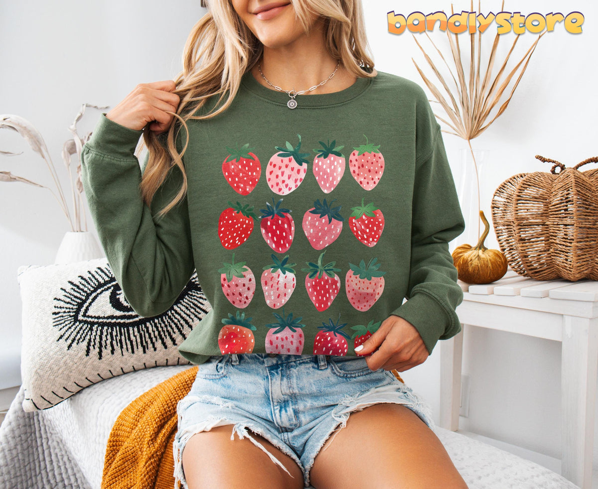 Cute Strawberry Sweatshirt in military green on a model seated indoors amid pillows, dried plants and wicker decor.