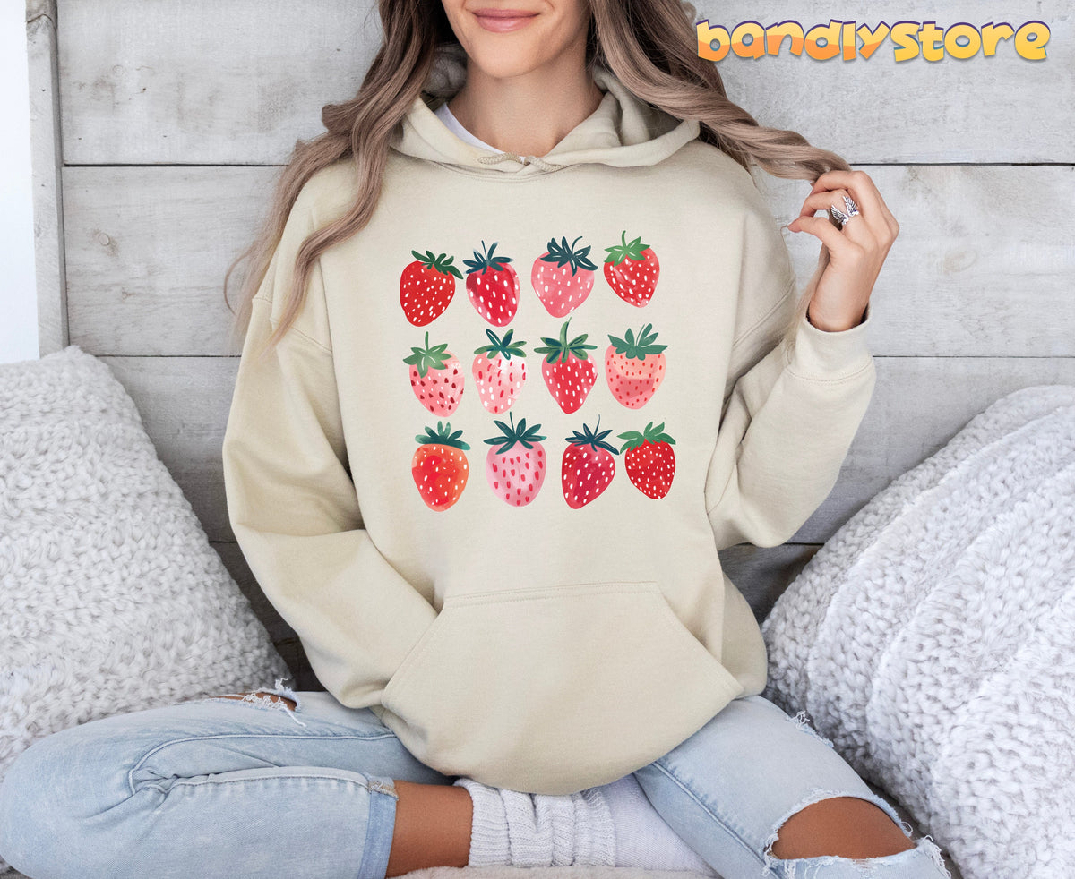 Cute Strawberry Hoodie in sand, worn front-facing by a seated person amid pillows and wooden wall.