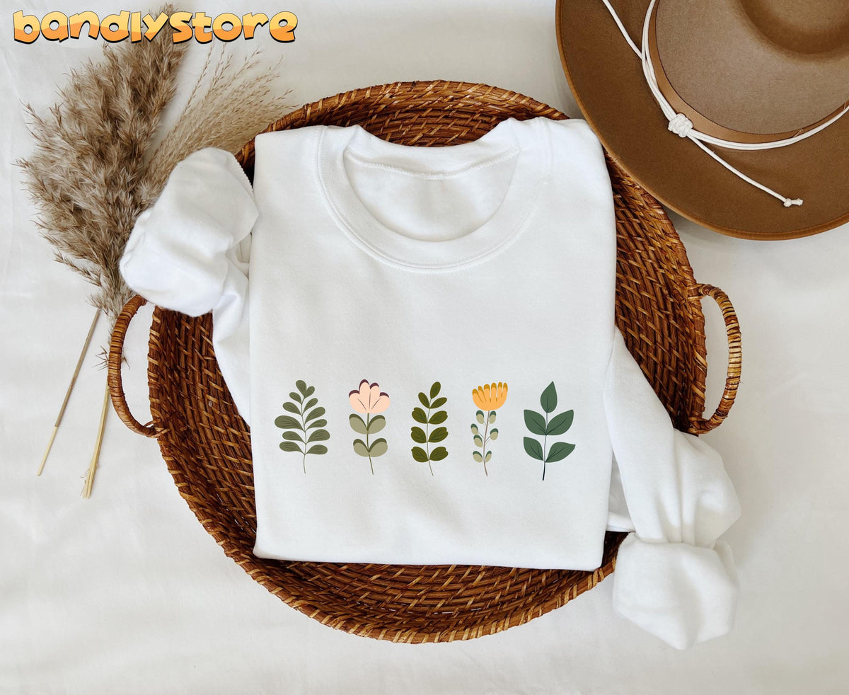 Cute Wildflowers Sweatshirt in white folded in a wicker basket beside dried grass and a hat.