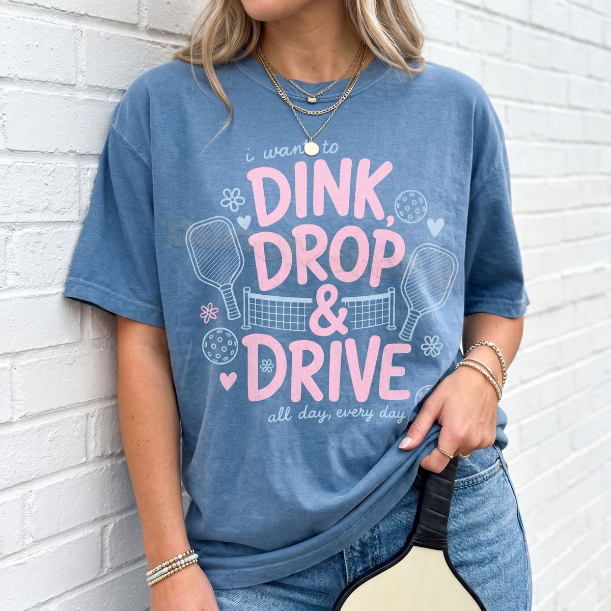 Dink Drop And Drive T-Shirt in Blue Jean, front view on person leaning against white brick wall holding pickleball paddle.