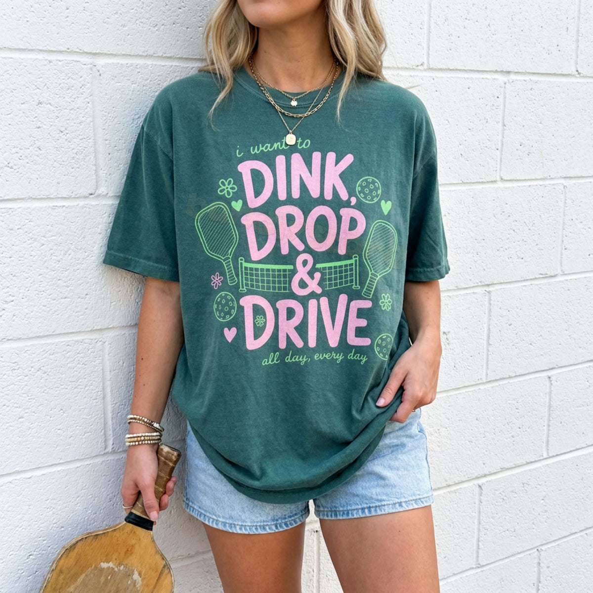 Dink Drop And Drive T-Shirt in Blue Spruce, worn front-facing by a woman holding a paddle against a white brick wall.