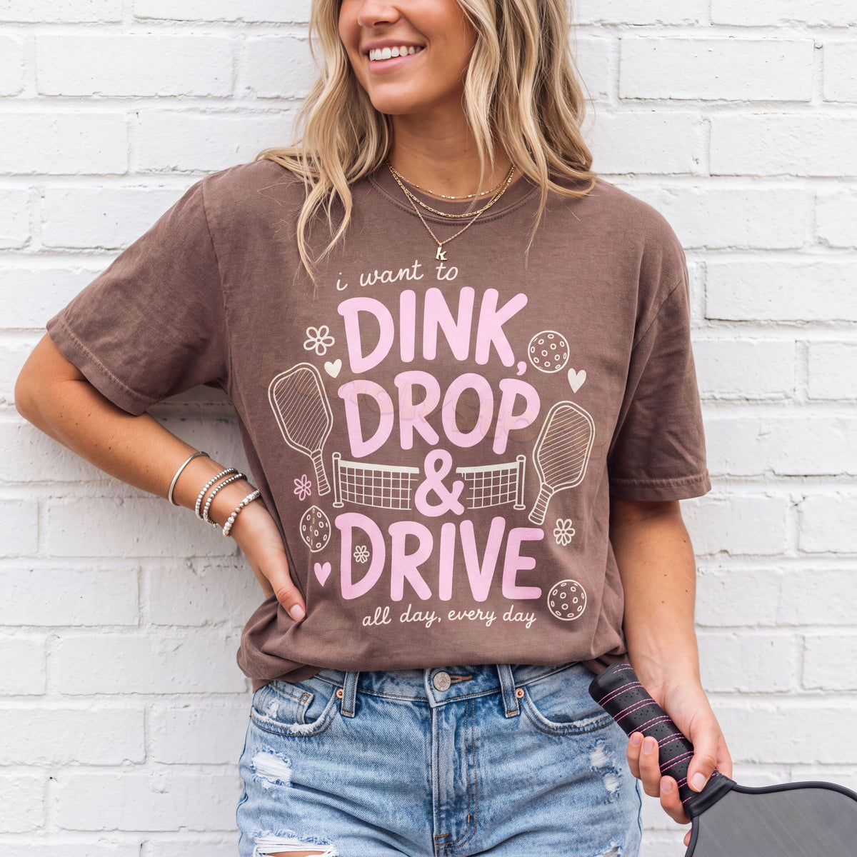 Dink Drop And Drive T-Shirt in espresso on a woman against a white brick wall, holding a pickleball paddle.