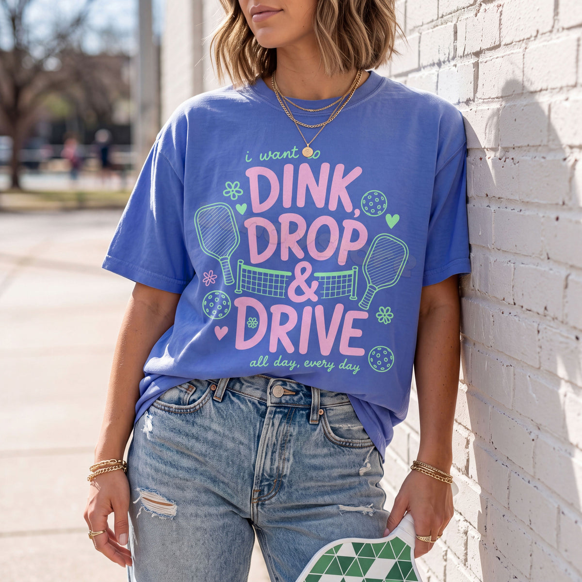 Dink Drop And Drive T-Shirt in Flo Blue worn front-facing against a brick wall outdoors with a paddle held.