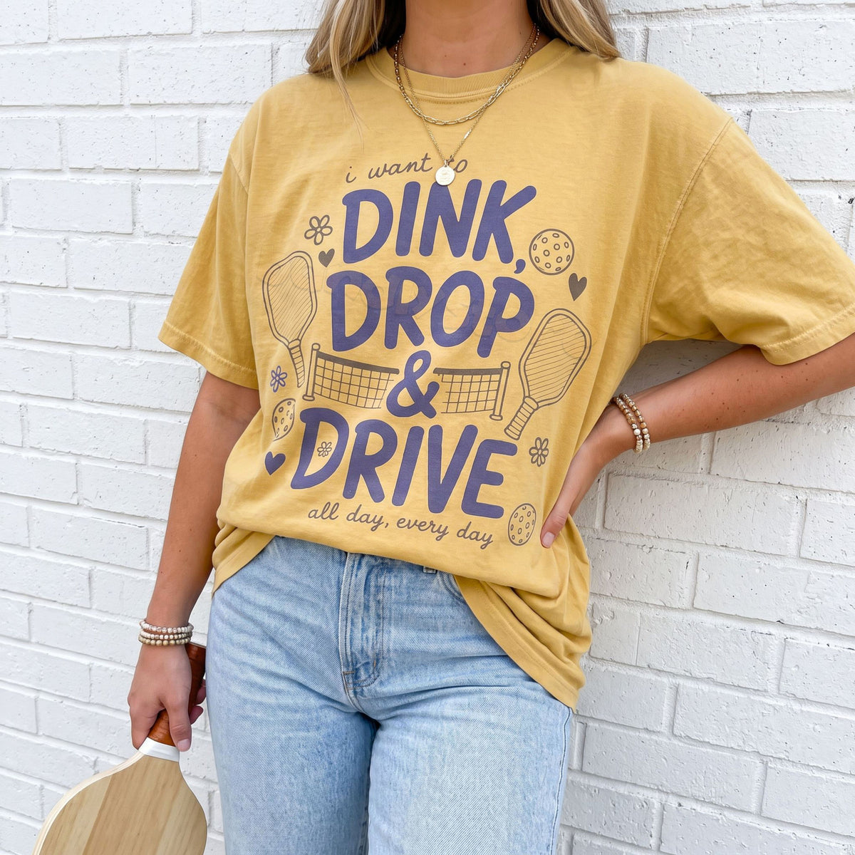 Dink Drop And Drive T-Shirt in mustard on a person against a white brick wall holding a paddle.