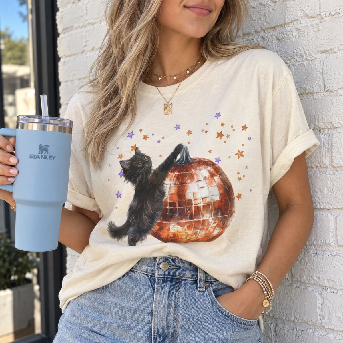 Black Cat Disco Ball Pumpkin T-Shirt in ivory, worn by a woman holding a blue tumbler against a white brick wall.
