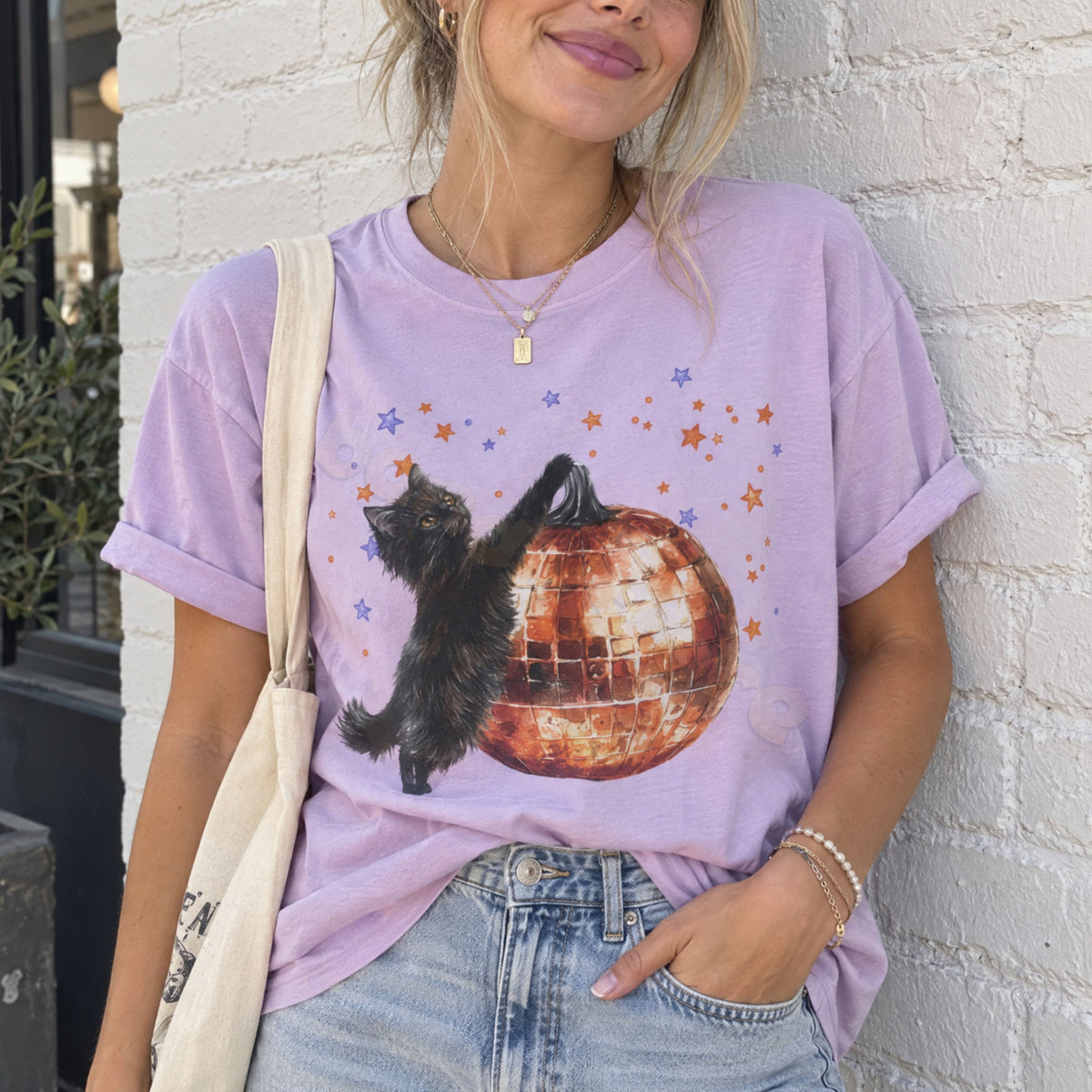 Black Cat Disco Ball Pumpkin T-Shirt in orchid on woman against white brick wall with tote bag.