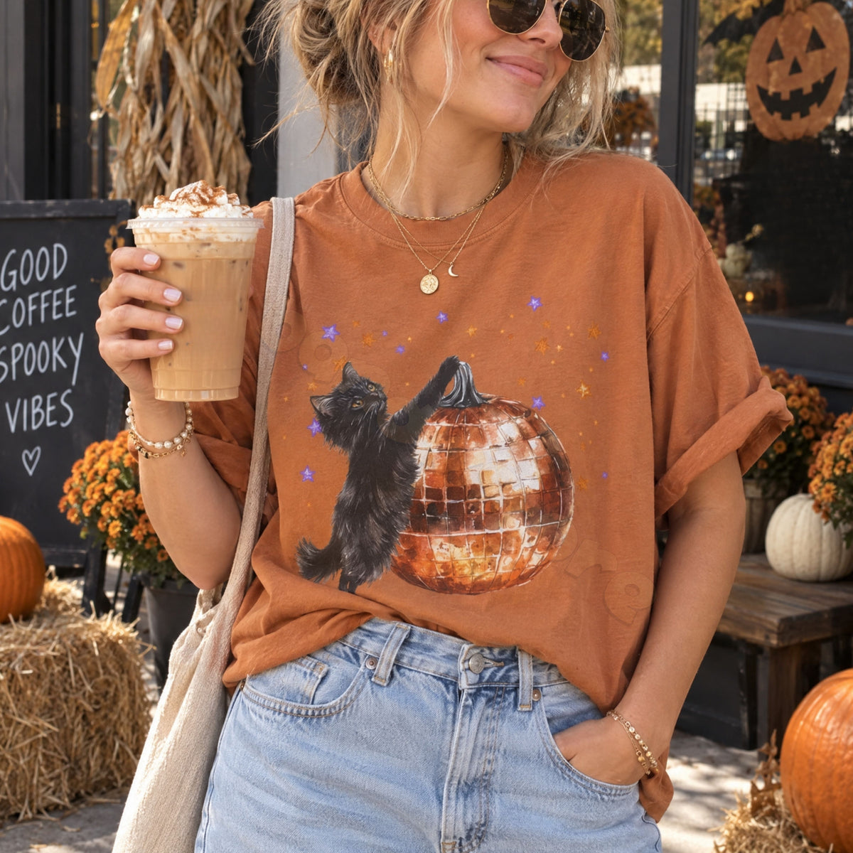 Black Cat Disco Ball Pumpkin T-Shirt in Yam, worn front-facing outdoors amid pumpkins, hay bales and a coffee sign.