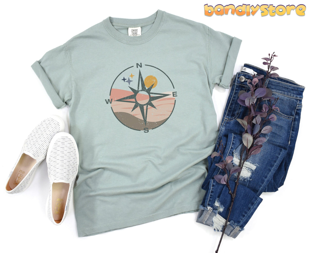 Distressed Compass T-Shirt in light green laid flat beside white slip-on shoes and blue jeans with a purple branch.