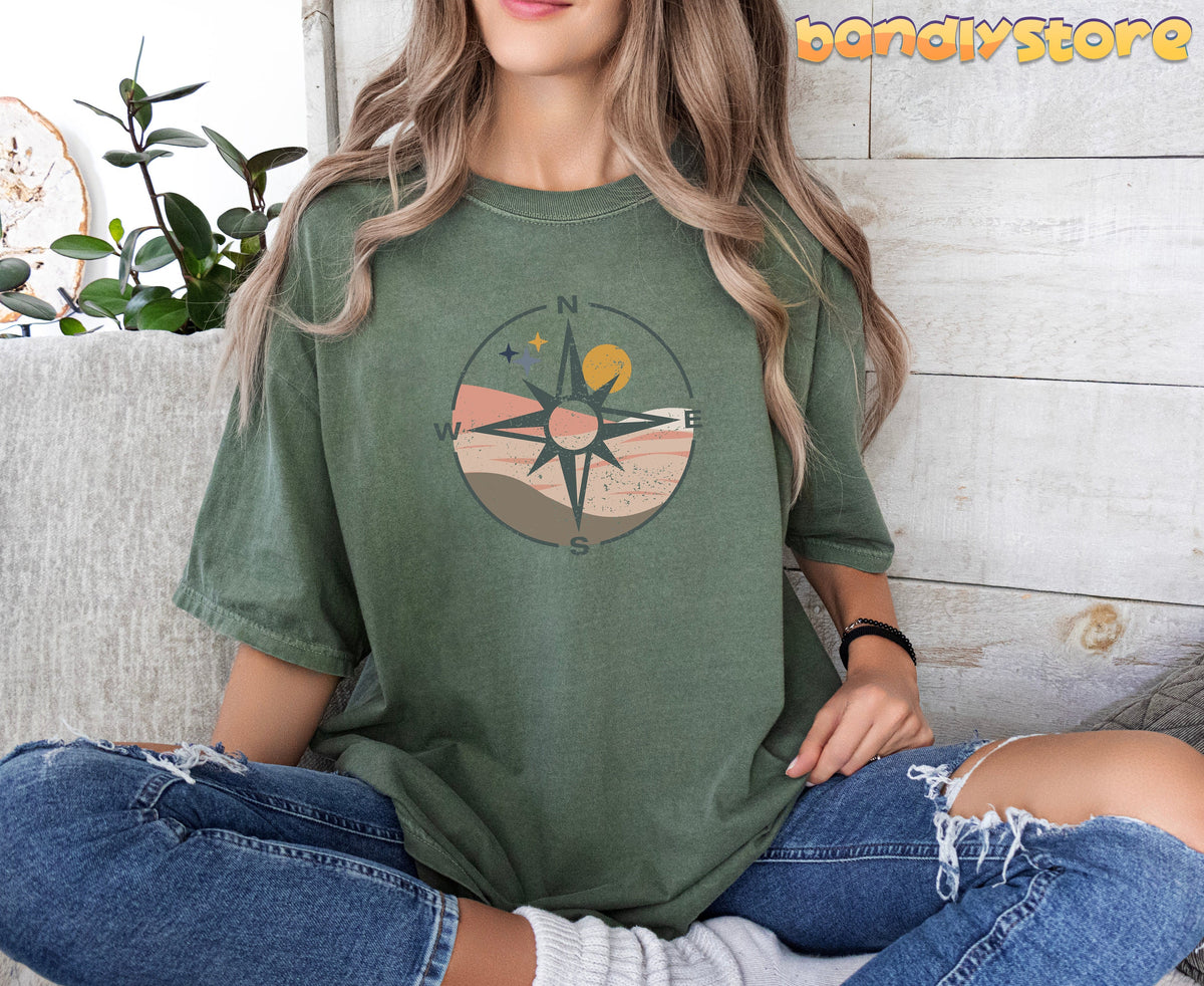 Distressed Compass T-Shirt in moss on a seated woman by a plant against a wood wall.