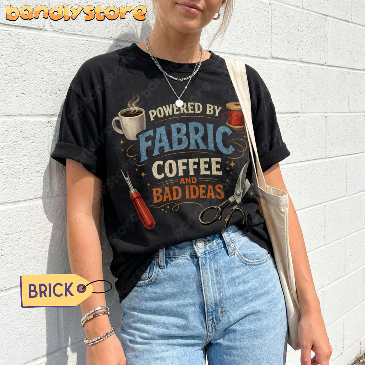 Powered By Fabric, Coffee And Bad Ideas T-Shirt in black on a person against white brick wall with jeans and tote bag.