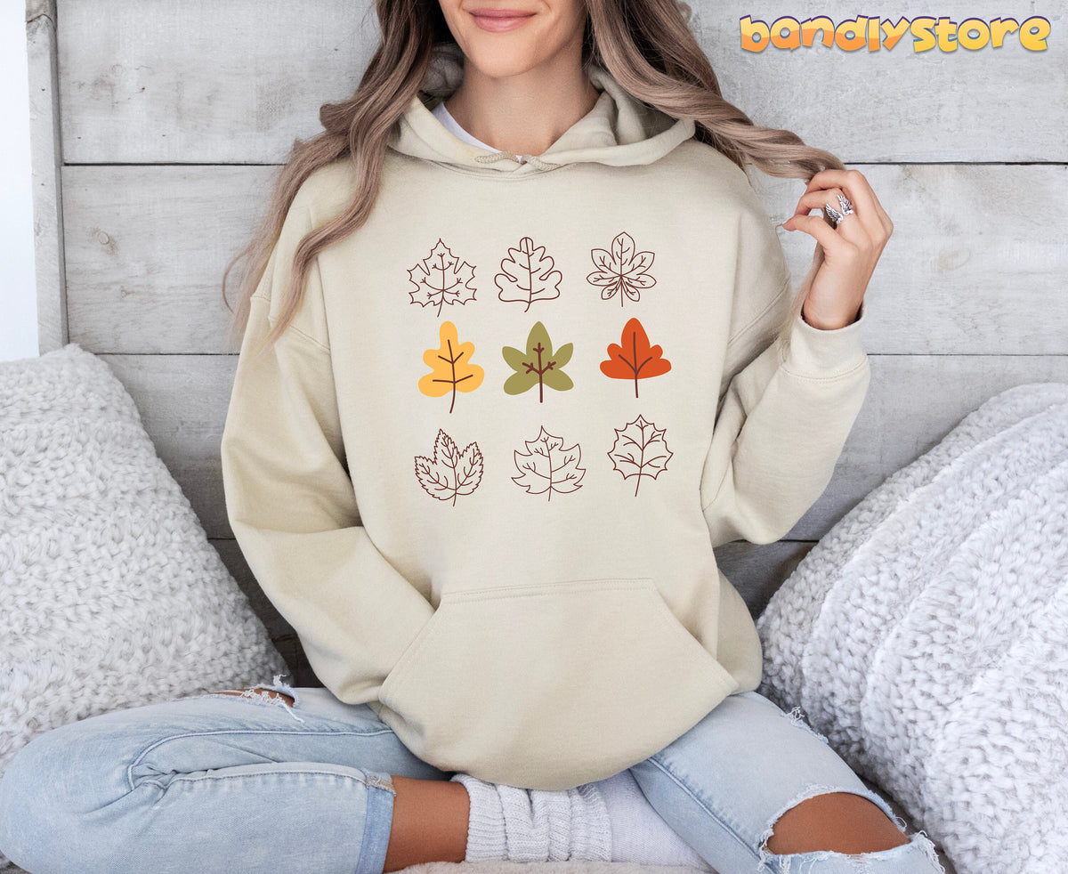 Fall Leaves Hoodie in sand, modelled front-on while seated amid white knit pillows on wood.