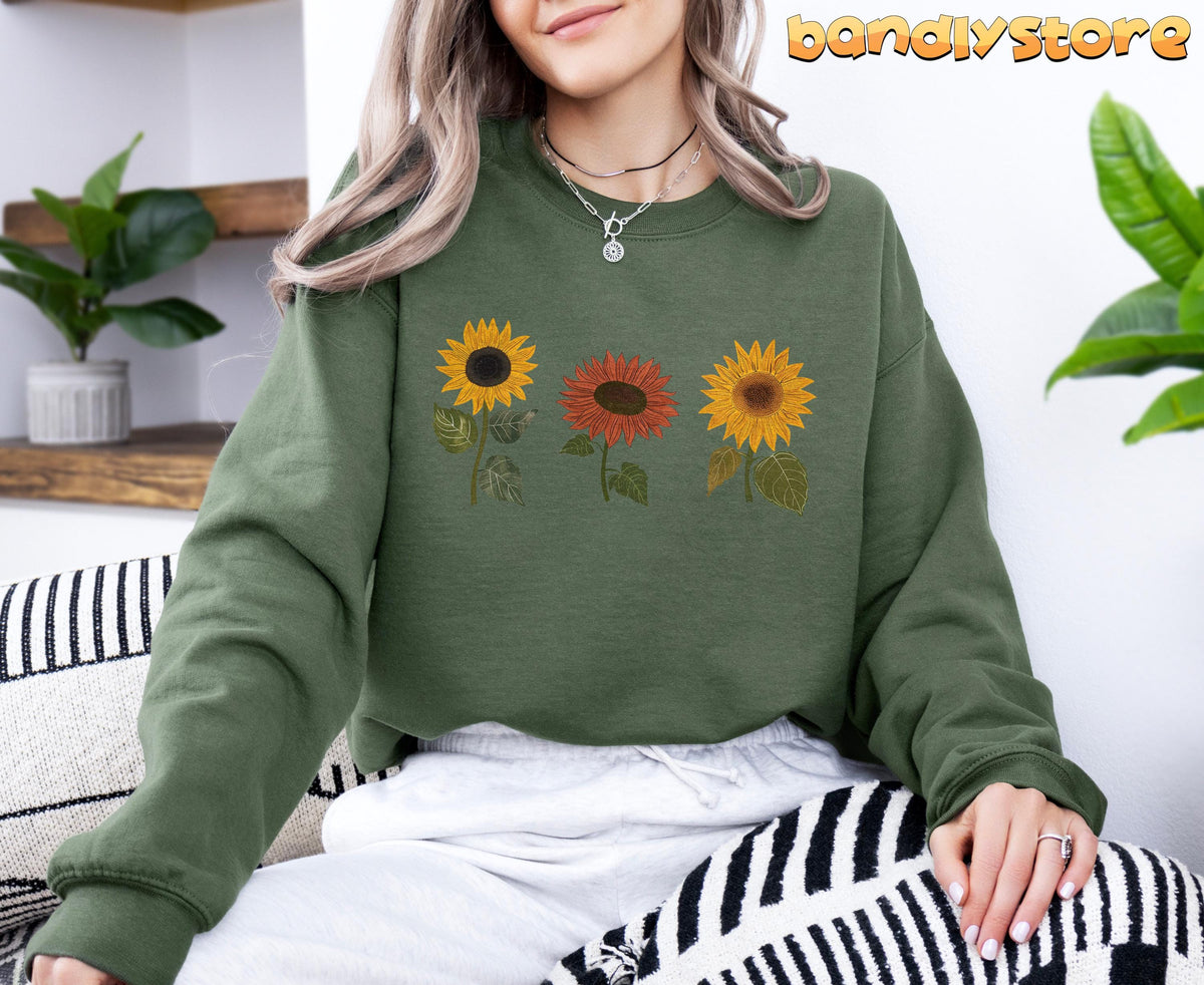 Fall Sunflower Sweatshirt in military green, worn seated on a patterned couch beside shelves and plants.
