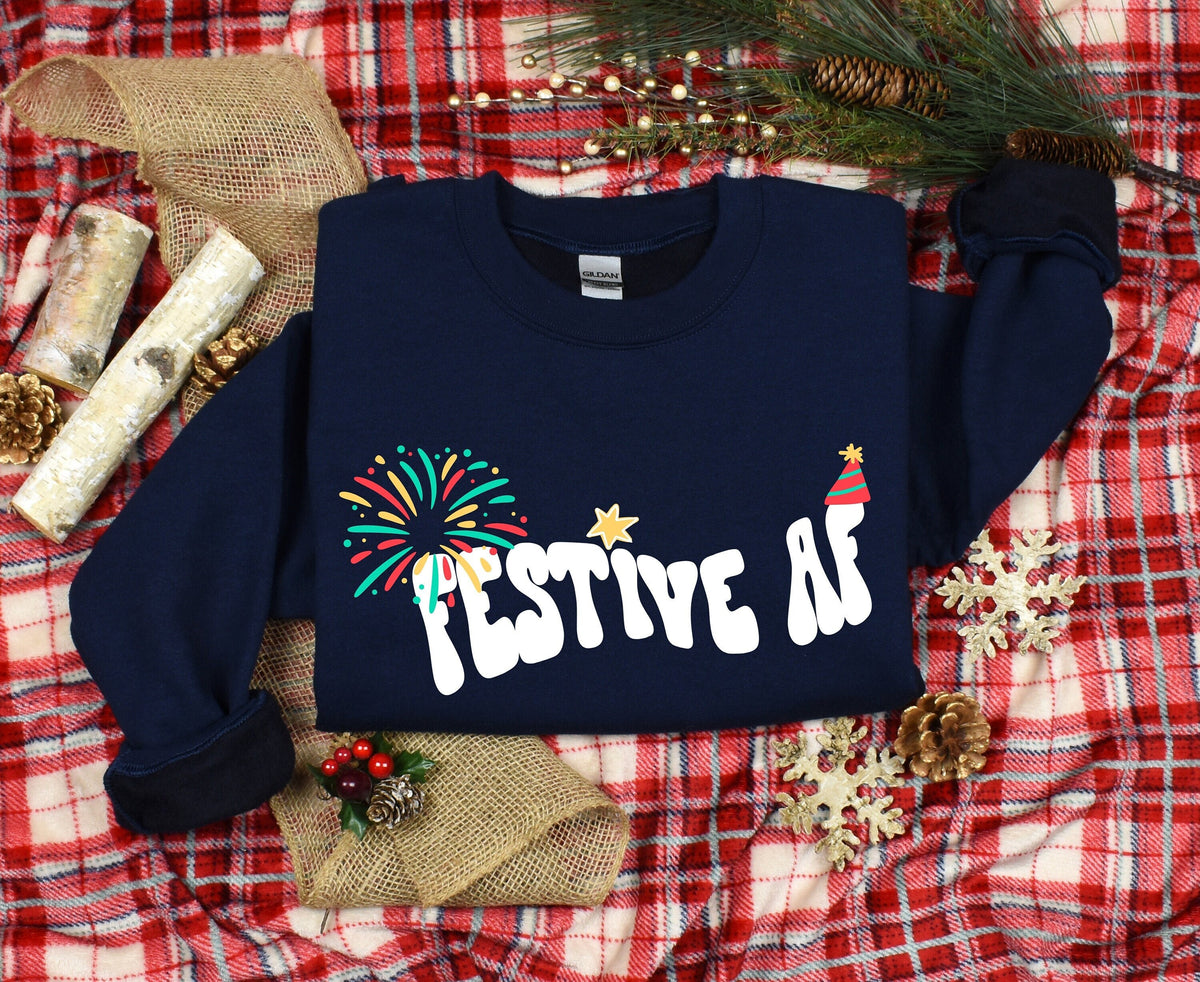 Festive AF Sweatshirt in navy laid flat on red plaid amid pinecones, burlap, birch logs and greenery.