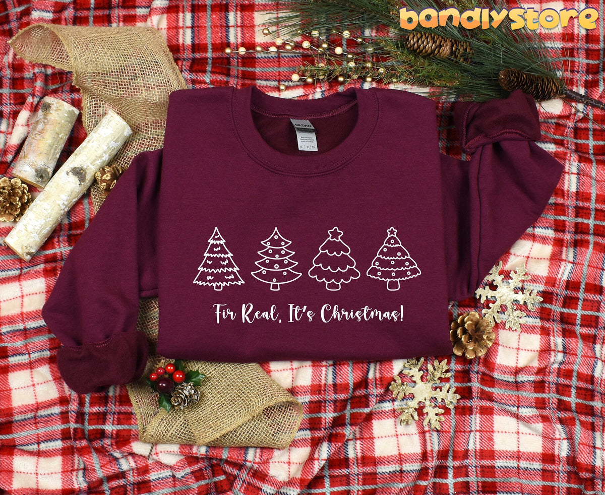 Fir Real It's Christmas Sweatshirt in maroon laid flat on plaid amid pinecones, birch logs, burlap and snowflake decorations.