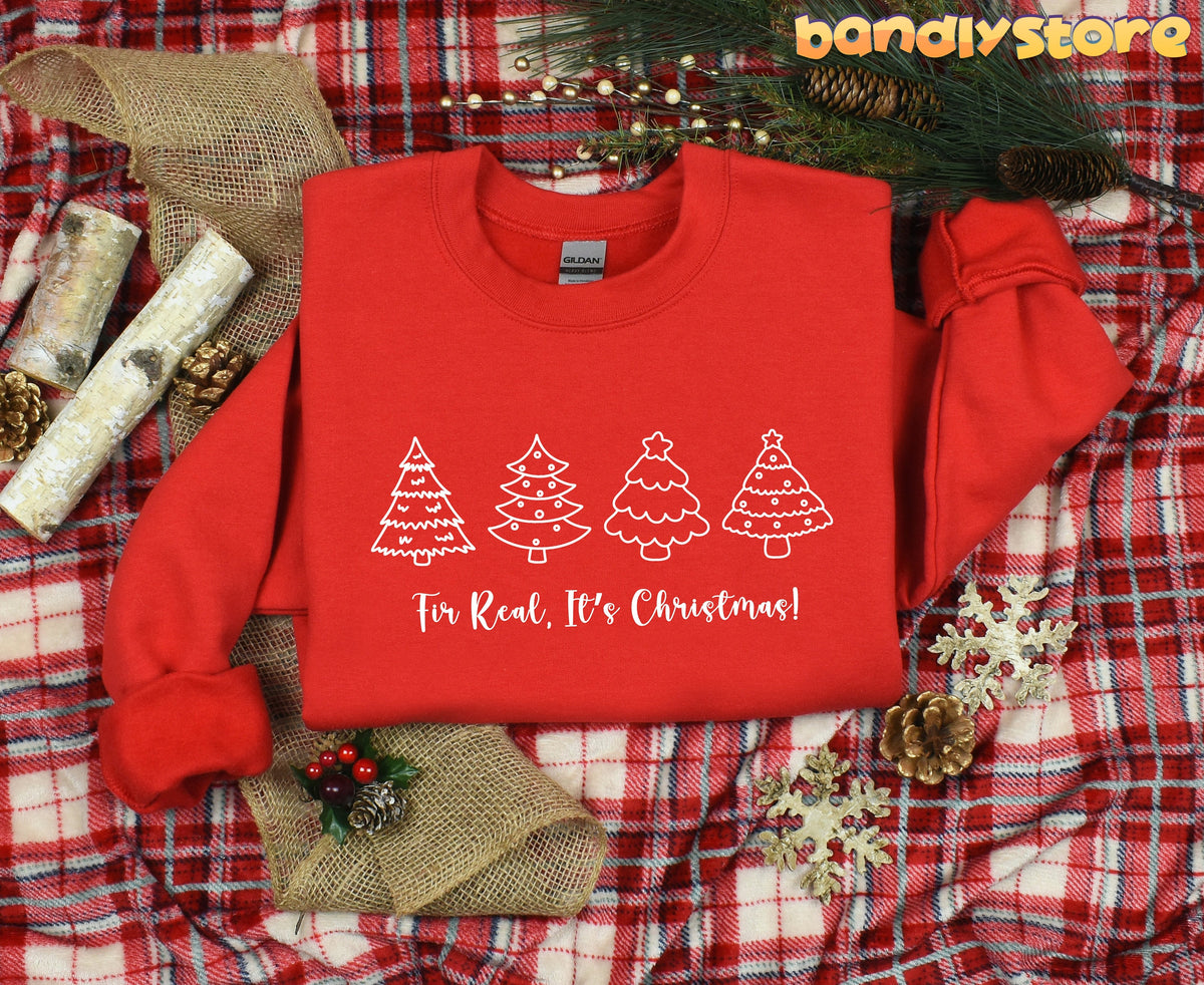 Fir Real It's Christmas Sweatshirt in red, top view on plaid amid pine greenery, cones, logs and snowflakes.