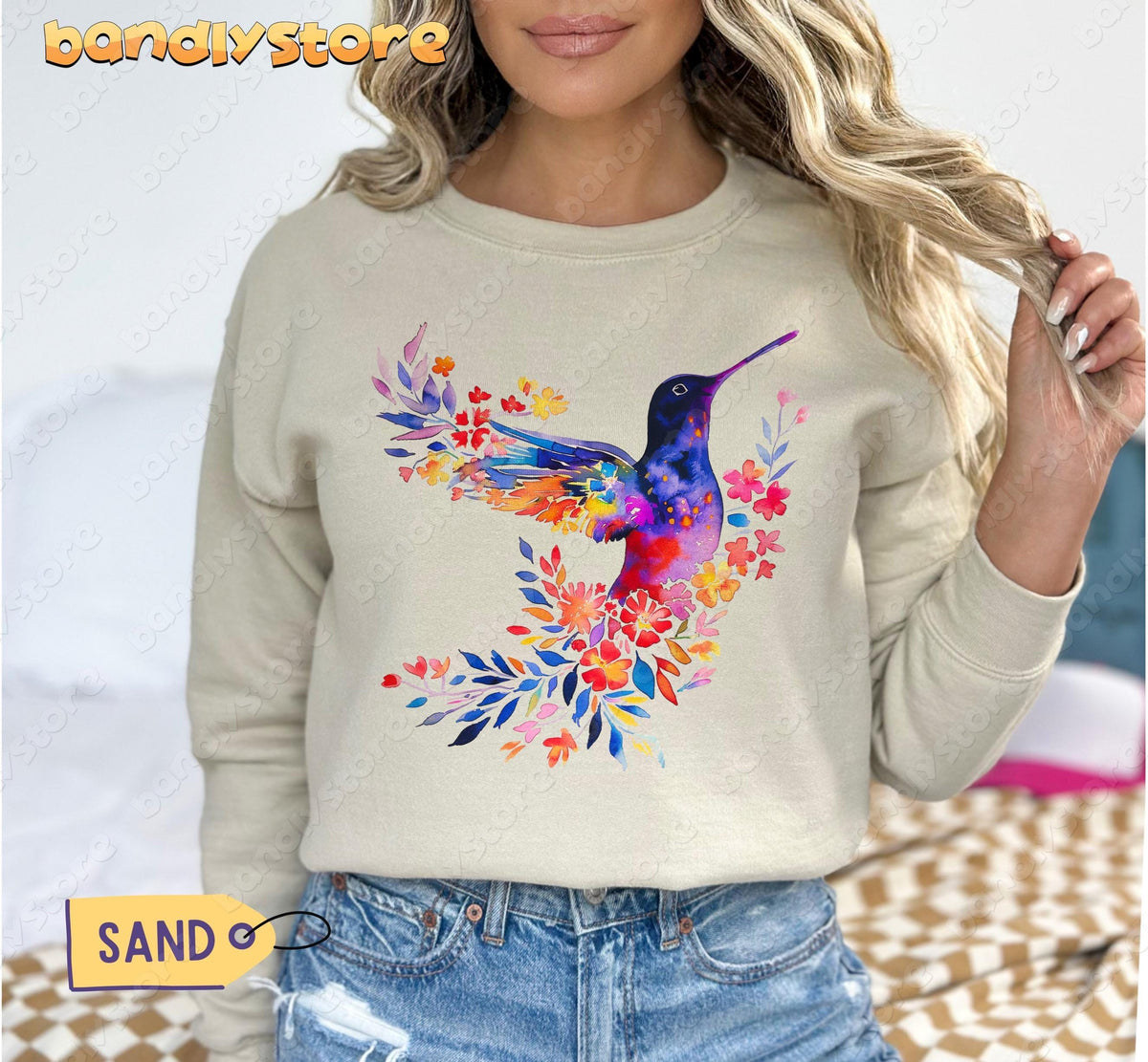 Floral Hummingbird Sweatshirt in sand, front view on a person by a bed with checkered bedding.