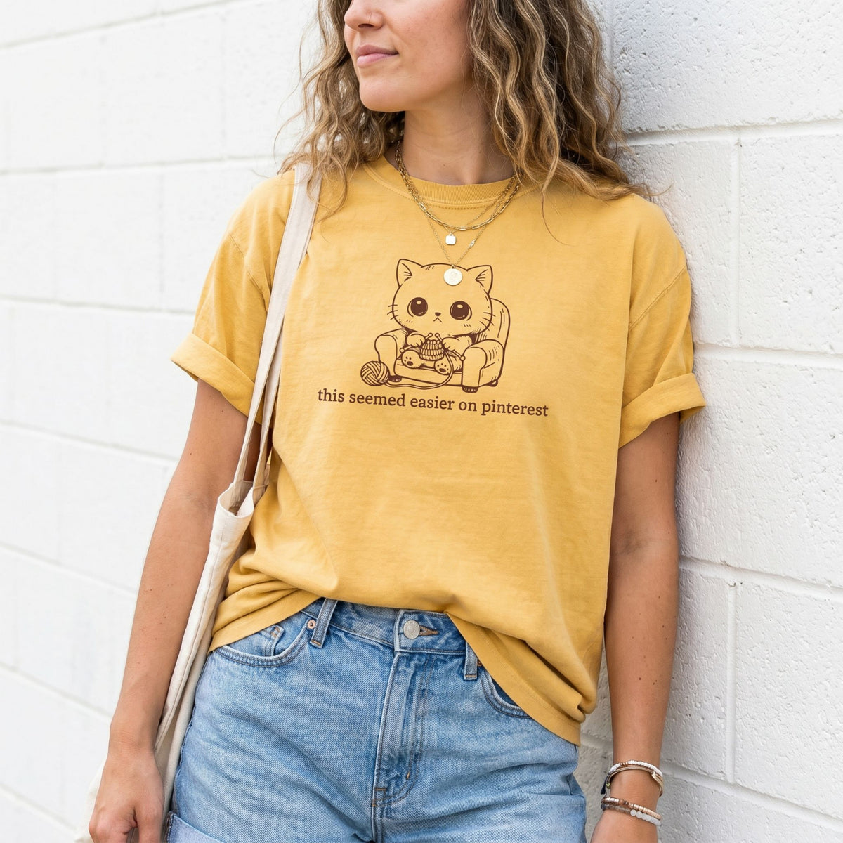Funny Knitting Cat T-Shirt in mustard worn by a woman leaning on a white brick wall with tote bag.