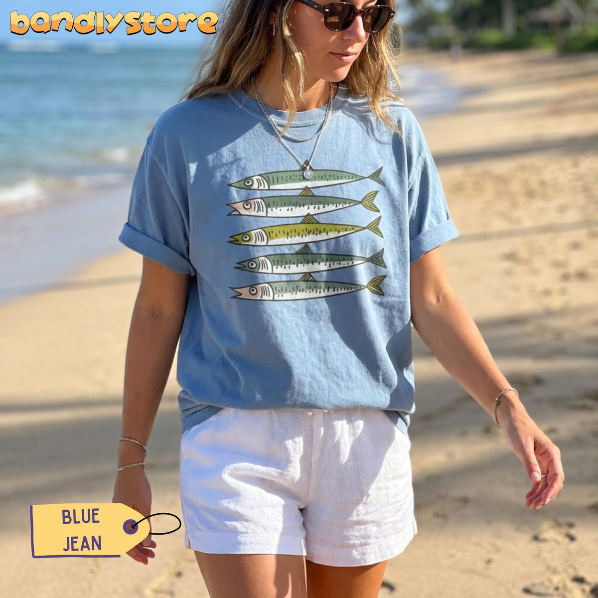 Funny Sardines T-Shirt in Blue Jean on a woman standing on a sunny beach by the ocean.