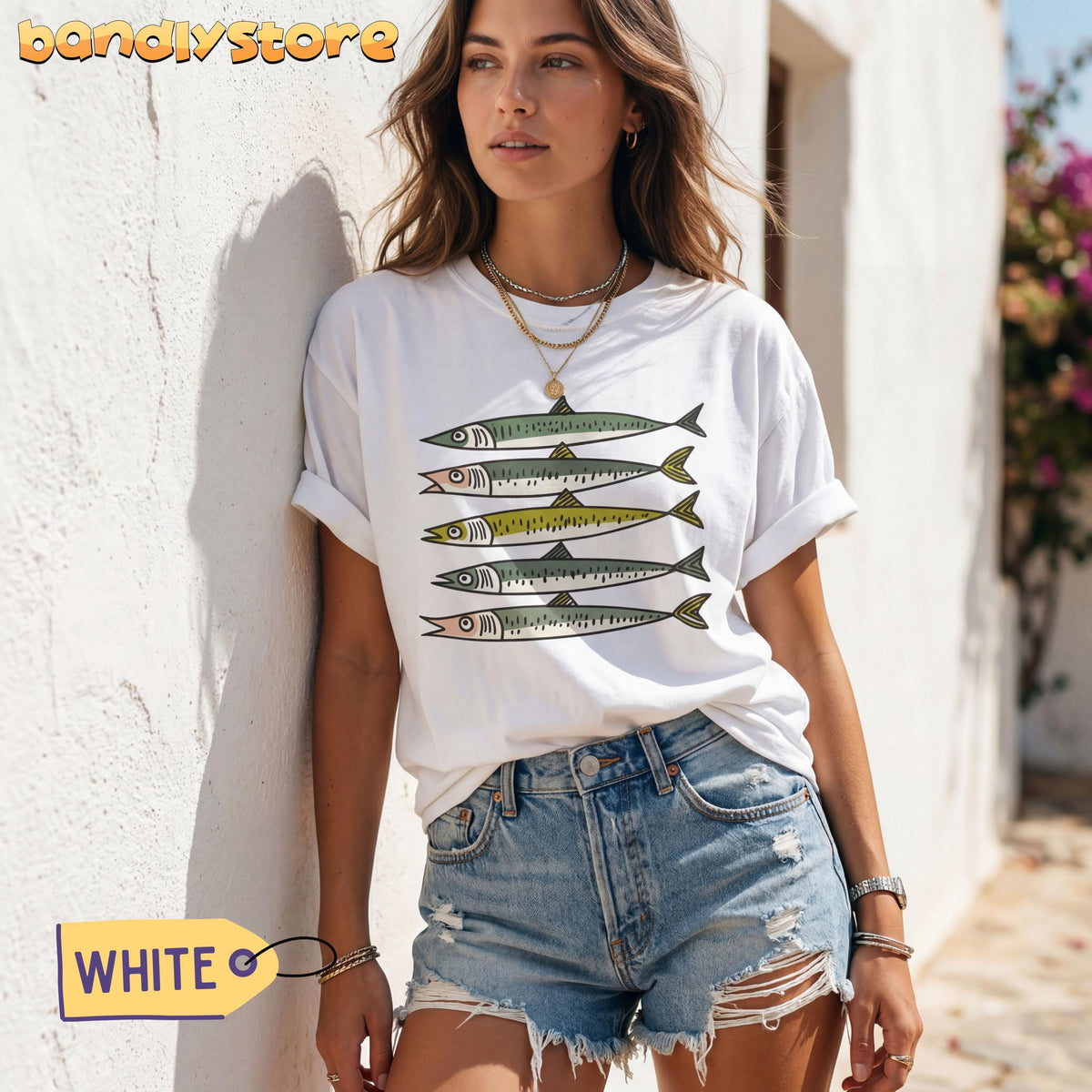 Funny Sardines T-Shirt in white on a woman leaning against a sunlit outdoor wall near flowering plants.