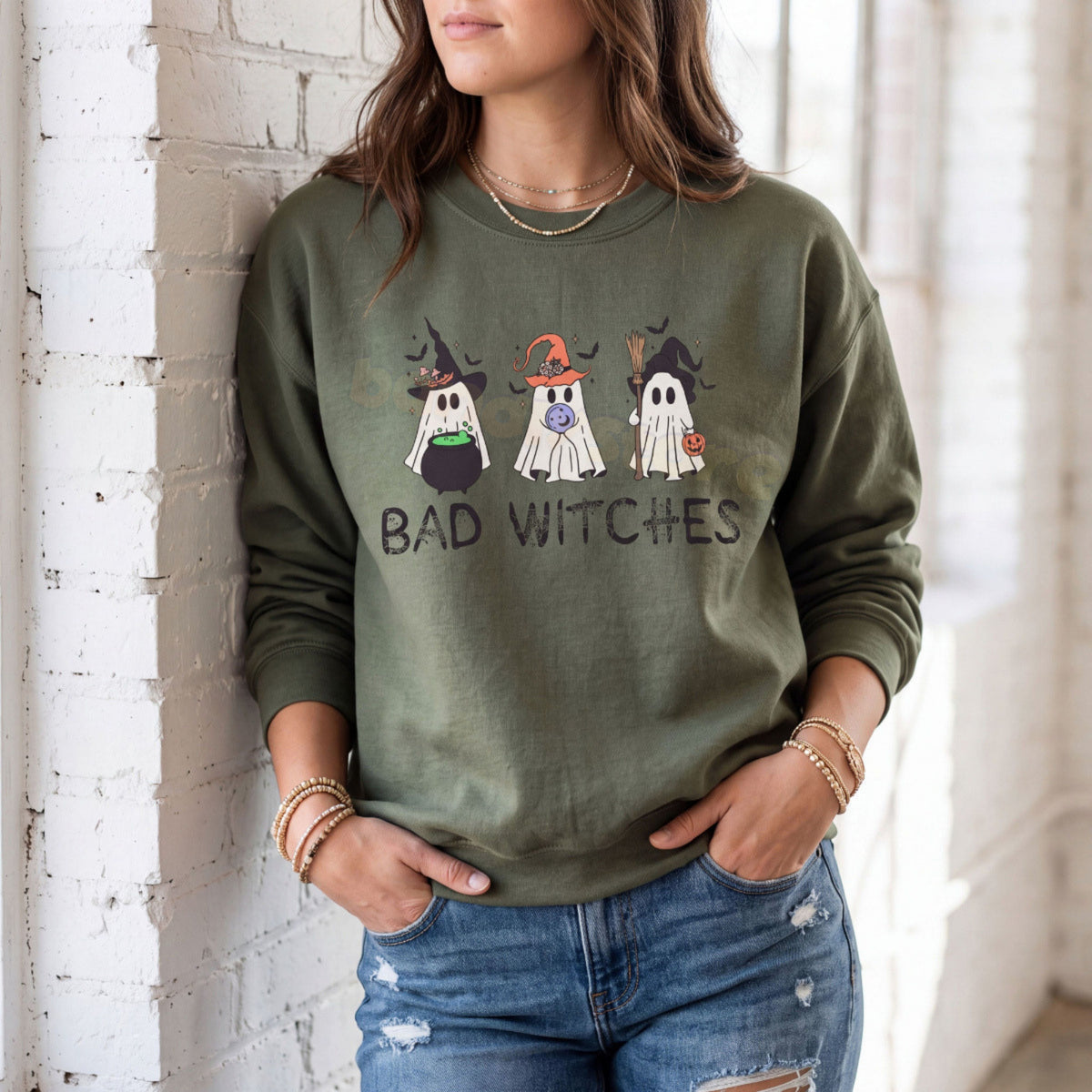 Ghost Bad Witches Sweatshirt in Military Green, woman leaning on white brick wall by window with hands in ripped jeans pockets.