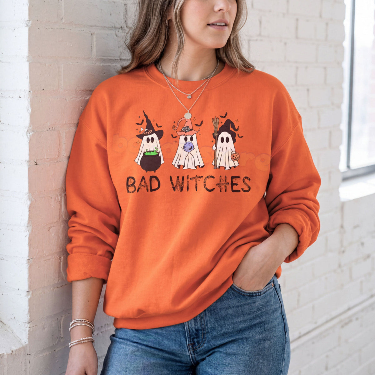 Ghost Bad Witches Sweatshirt in orange on a woman leaning against a white brick wall by a window.