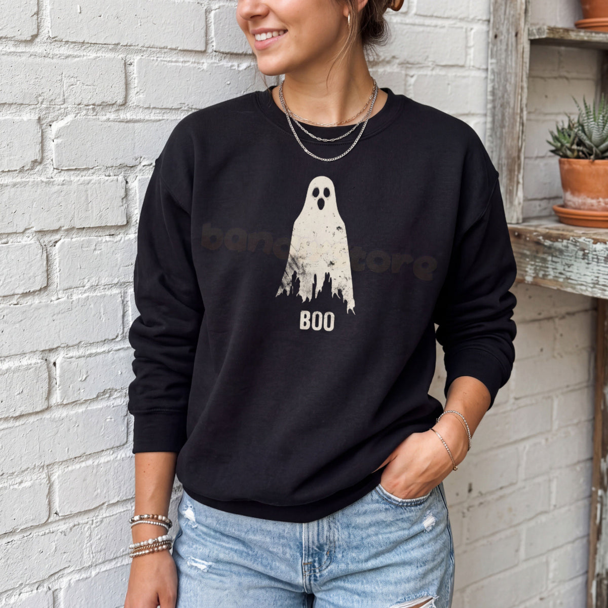 Grungy Ghost Sweatshirt in black on a person against white brick wall beside wooden shelves with plants.
